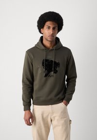 Haut à capuche vert kaki avec un design graphique sur le devant représentant une tête de lion. Il possède une capuche avec cordon de serrage et des poignets côtelés. Porté avec un pantalon beige clair.