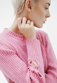 Jersey de punto acanalado rosa con cuello redondo y detalles cortados en las mangas, combinado con pendientes de aro dorados y un anillo.
