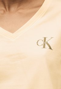 Κοντινό πλάνο ενός μπεζ πουκαμίσου με V-λαιμόκοψη και μικρό λογότυπο Calvin Klein "cK" τυπωμένο στο αριστερό στήθος.