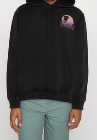 Svart huvtröja med en framficka, prydd med en grafisk palmträd i lila och gult. Mjuk textur och justerbara snoddar på huvan.