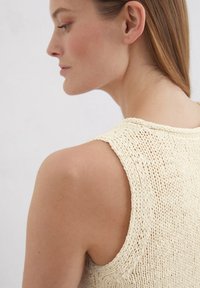 Top senza maniche lavorato a maglia in un morbido color crema con un design aperto e texturizzato. Presenta un ampio scollo e spalle leggermente arrotondate.