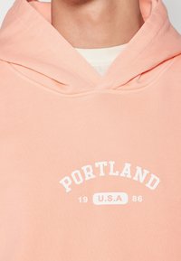 Pfirsichfarbener Baumwoll-Hoodie mit einem großen weißen "PORTLAND"-Grafik und "19 U.S.A 86" darunter. Verfügt über eine Kordelzugkapuze und gerippte Bündchen.