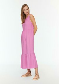 Maxikleid aus strukturiertem, hellrosa Stoff, ärmellos mit eng anliegendem Oberteil und gestuften Rüschensaum. Kombiniert mit neutralen Strandsandalen.