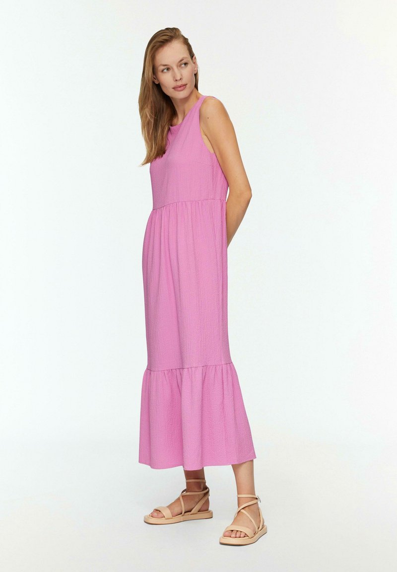 Maxikleid aus strukturiertem, hellrosa Stoff, ärmellos mit eng anliegendem Oberteil und gestuften Rüschensaum. Kombiniert mit neutralen Strandsandalen.