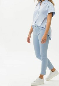 Pantaloni attillati azzurri, con chiusura a bottone e cerniera, abbinati a una camicia a maniche corte a righe azzurre. Sneakers bianche completano l'outfit.