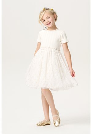 Weißes Kleid mit geripptem Oberteil und Tüllrock, verziert mit goldenen Punkten; kombiniert mit goldenen Ballettschuhen. Das Mädchen steht mit den Händen auf den Hüften und lächelt.