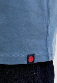 Blauer Strickstoffsaum mit einem kleinen schwarzen Etikett mit rotem Kreis, über einer dunkelblauen Jeansnaht.