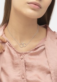 Collana d'argento con pendenti circolari interconnessi, su una delicata catena, indossata sopra una morbida camicia rosa con bottoni e una leggera lucentezza.