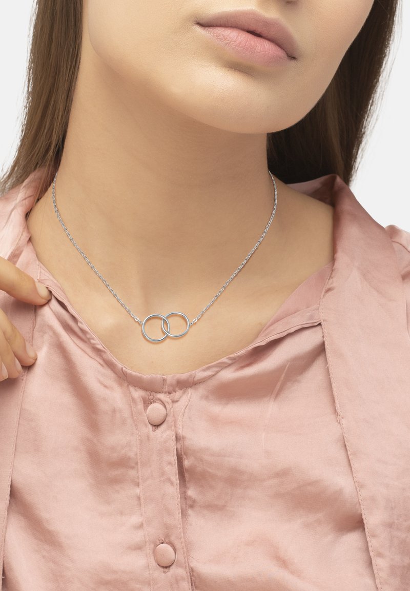 Collana d'argento con pendenti circolari interconnessi, su una delicata catena, indossata sopra una morbida camicia rosa con bottoni e una leggera lucentezza.