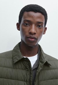 Olive green puffer jacket with a structured collar, textured fabric, and snap-button accents, worn over a gray sweater and white top.