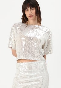 Top corto in paillettes argento con maniche corte, caratterizzato da una vestibilità morbida e una texture lucida, abbinato a una gonna coordinata in paillettes.