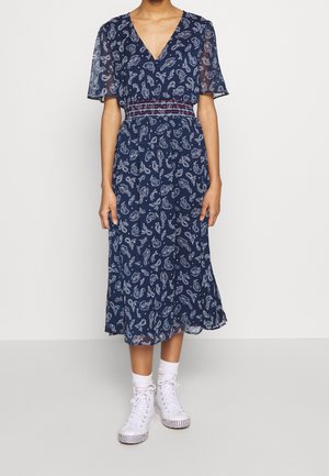 Person trägt ein midilanges Marineblaues Paisley-Kleid mit transparenten kurzen Ärmeln, einer gerafften Taille, weißen Socken und weißen High-Top-Sneakern.