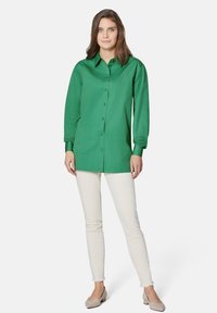 Grüne Knopfhemd mit Kragen und langen Ärmeln, kombiniert mit weißen Hosen und hellgrauen Ballerinas. Glatte, einfarbige Textur.