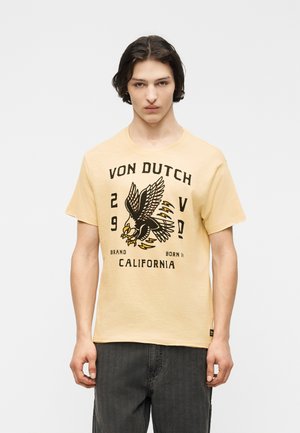 Noor mees tumedate juustega, kandes beeži Von Duchi T-särki, millel on suur kotkas graafika, ja musti pükse, seistes tavalise tausta ees.