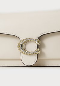Beige leather handbag with silver and yellow crystal embellishment on a prominent clasp, featuring clean lines and contrasting black stitching.