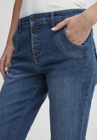 Sinine denim seelik kõrgema vöökohaga, millel on ees viis nuppu, küljetaskud ja nähtavad õmbluse aktsendid. Sile tekstuur.