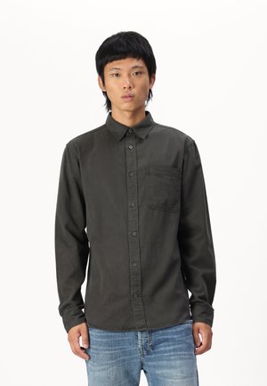 Chemise boutonnée grise avec une poche, manches longues et col classique. Le tissu semble lisse avec un léger éclat. Associée avec un jean.