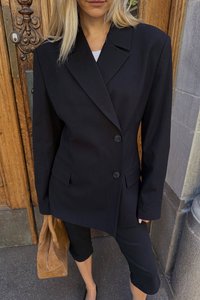 Zwarte oversized blazer met twee knopen, scherpe revers en voorkeurszakken, gedragen met zwarte aansluitende broek en een beige tas. Op de achtergrond is een houten deur te zien.