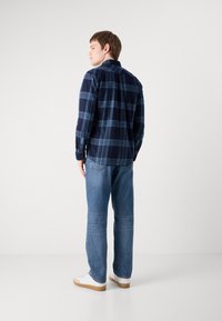 Camisa de xadrez azul marinho com riscas azuis claras, com colarinho abotoadinho e mangas longas. Usada com jeans de ganga azul clara e ténis brancos.