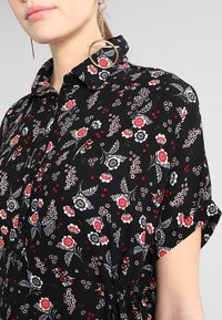 Schwarze Bluse mit floralen Mustern in Rot und Weiß, einem kragenförmigen Halsausschnitt, kurzen Ärmeln und Raffung an der Taille.