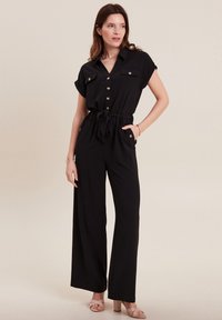 Zwarte jumpsuit met korte mouwen, knoopsluiting aan de voorkant, tailleband en twee borstzakken. Gemaakt van een gladde stof met wijde broekspijpen.