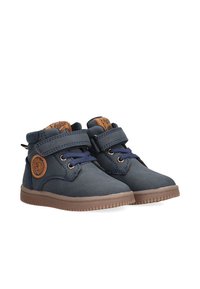 Zapatillas de lona azul con suelas de goma marrón, con una tira de velcro, cordones redondos y un logo circular en el lateral.