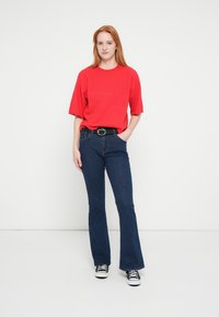 Top rojo de manga corta, vaqueros acampanados azules con cinco bolsillos, cinturón negro con hebilla plateada y zapatillas negras y blancas. Fondo liso.