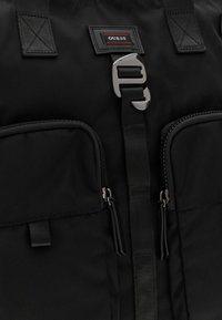 Zaino nero in nylon con una cinghia centrale dotata di fibbia metallica, due tasche con zip e un'etichetta di marca con dettagli rossi.