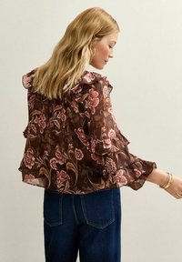 Bruine bloemenblouse met gerimpelde schouders en lange mouwen. Heeft een doorschijnende stof met roze, rode en crème bloemenpatronen.