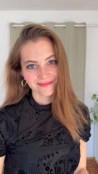 Jovem sorridente com cabelo castanho claro longo, olhos azuis, usando brincos de argola dourados e um top preto com padrão, em pé numa divisão interior.