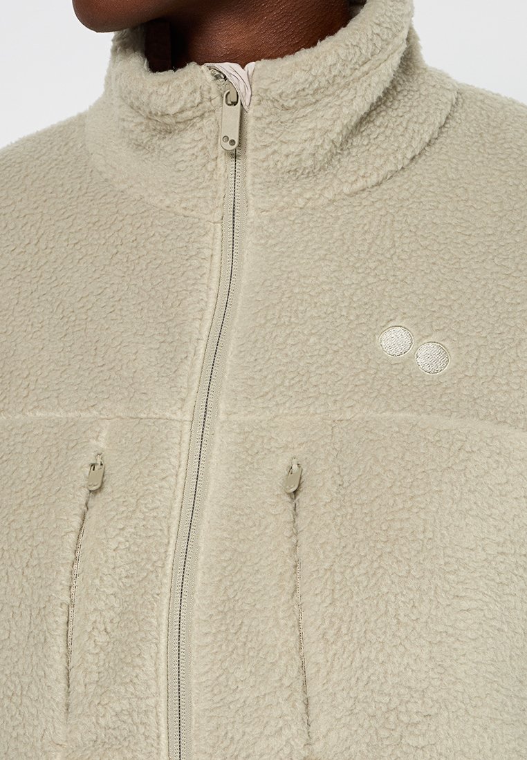 Veste en polaire beige avec un col haut, une fermeture éclair à l'avant et deux poches latérales. Présente un petit logo dans le coin supérieur droit et une texture douce.