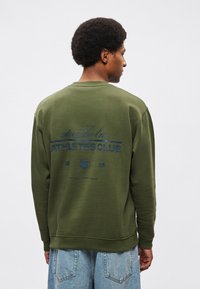 Sudadera de algodón verde oliva con un gráfico azul en la espalda que dice "stockholm ATHLETES CLUB 2015", con puños y dobladillo acanalados.
