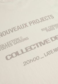 Camiseta de algodón en color crema con texto impreso en gris. Presenta frases como "NOUVEAUX PROJECTS" y "COLLECTIVE DE," con una textura suave.