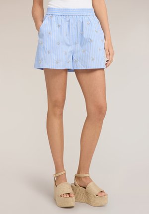 Kobieta ubrana w jasnoniebieskie szorty w paski z małym kwiatowym haftem i beżowe sandały na platformie, stojąca na jednolitym tle.