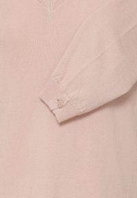 Pull rose clair en tricot avec un col en V et une manche longue à manchette boutonnée. La texture est douce et lisse.