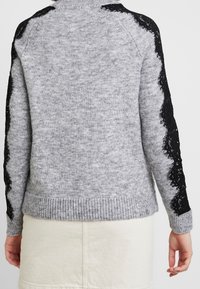 Maglione grigio lavorato a maglia con dettagli in pizzo nero sulle maniche, polsini e orlo a costine, con scollatura rotonda e finitura testurizzata.