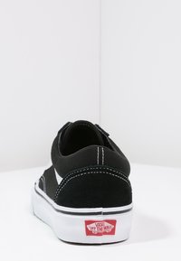 Baskets noires Vans avec semelle blanche, couture visible, logo rouge "Off The Wall" sur le talon, vues de dos sur une surface blanche avec un fond blanc.