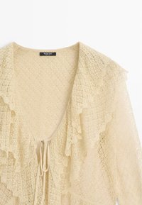 Cardigan beige lavorato a maglia con scollo a V profondo, bordi arricciati e chiusura con laccio sul davanti.