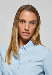 Ljusblå knapp-up skjorta i ett mjukt material med en liten broderad logotyp. Klassisk krage och standardmanschetter. Ren design.