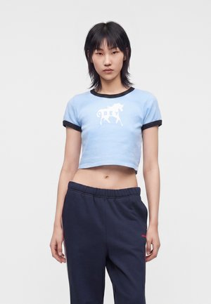 Osoba z czarnymi włosami ubrana w jasnoniebieski krótki t-shirt z białą grafiką zwierzęcia i ciemnoniebieskie spodnie dresowe, stojąca na białym tle.