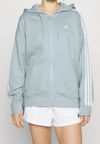Mikina na zip z měkké, světle modré látky. Má bílý logo Adidas a tři bílé pruhy podél rukávů. Volný střih.