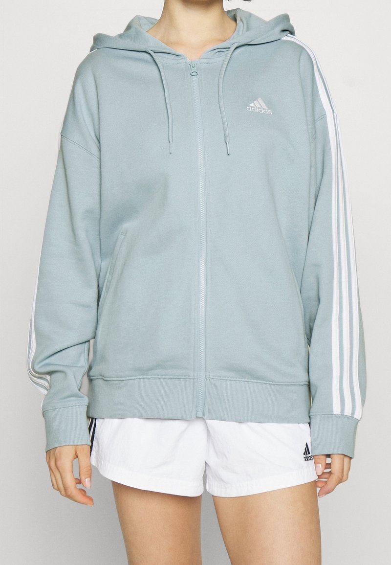 Mikina na zip z měkké, světle modré látky. Má bílý logo Adidas a tři bílé pruhy podél rukávů. Volný střih.