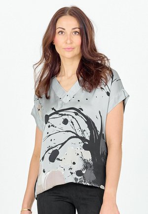 Ženska z dolgimi rjavimi lasmi, oblečena v svetlo sivo bluzo s črnim abstraktnim vzorcem iz črnil in črne hlače, stoji pred belim ozadjem.