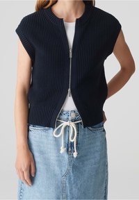 Kvinna i en ärmlös marinblå stickad zip-up väst över en vit topp, tillsammans med en ljusblå jeanskjol som är knuten med ett vitt repbälte.