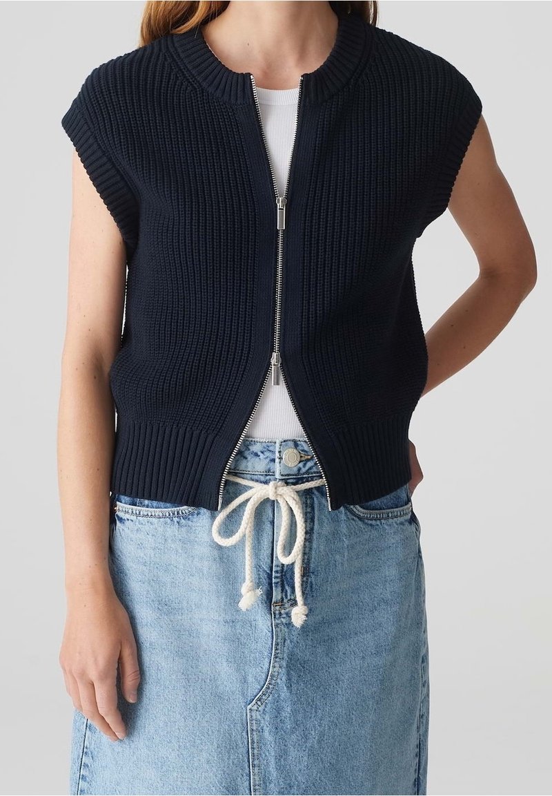 Kvinna i en ärmlös marinblå stickad zip-up väst över en vit topp, tillsammans med en ljusblå jeanskjol som är knuten med ett vitt repbälte.