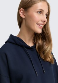 Giovane donna con lunghi capelli castano chiaro che sorride, indossando una felpa blu scuro con coulisse, su uno sfondo grigio chiaro.