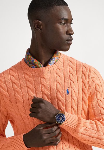 Cable-knit orange sweater, patterned shirt collar in green and blue, gold watch with blue face, worn on a model with dark skin.