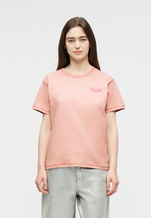 Jeune femme aux cheveux bruns lisses portant un t-shirt rose à manches courtes avec une petite rose brodée sur la poitrine et un pantalon de couleur claire.