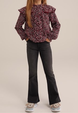 Florale Bluse in Dunkelrot mit weißen und rosa Blumen, Rüschensäumen und einem hohen Ausschnitt. Kombiniert mit schwarzen Schlagjeans und Turnschuhen.