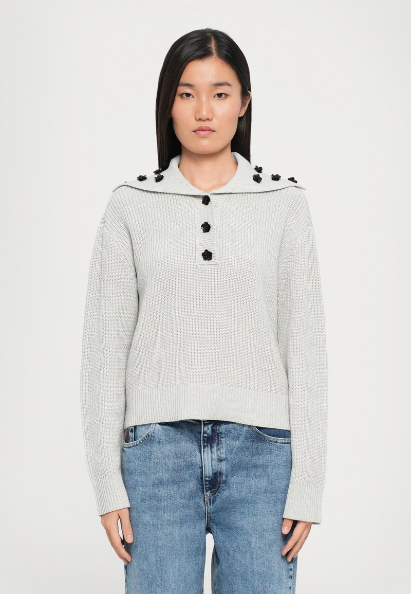 Pull en maille gris clair avec un col large, orné de boutons décoratifs noirs le long des épaules et de poignets et un ourlet côtelé.